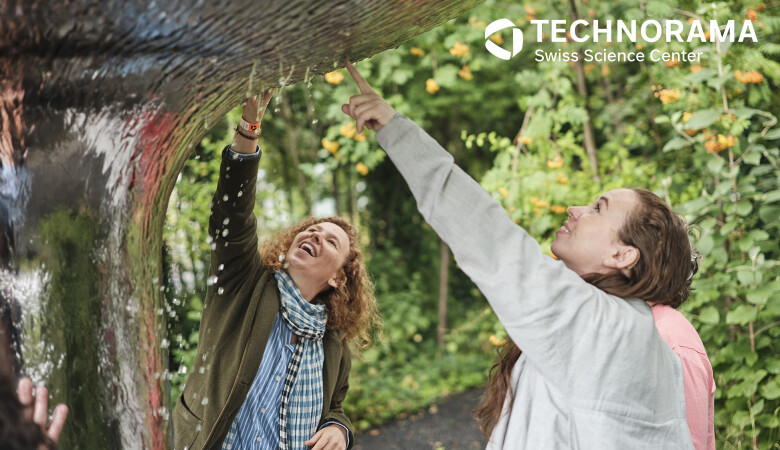 Zwei Frauen interagieren lachend mit einer Wasserinstallation im Aussenbereich des Swiss Science Center Technorama, umgeben von grüner Natur.